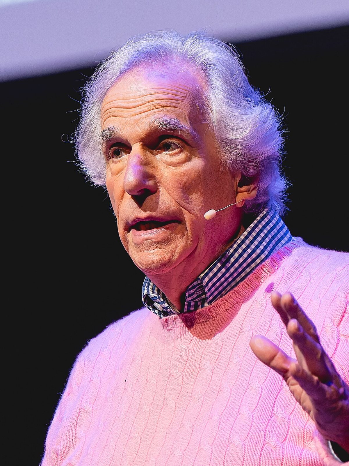 Henry Winkler, white-haired, in a pink sweater and checked shirt, speaking on stage with a headset microphone