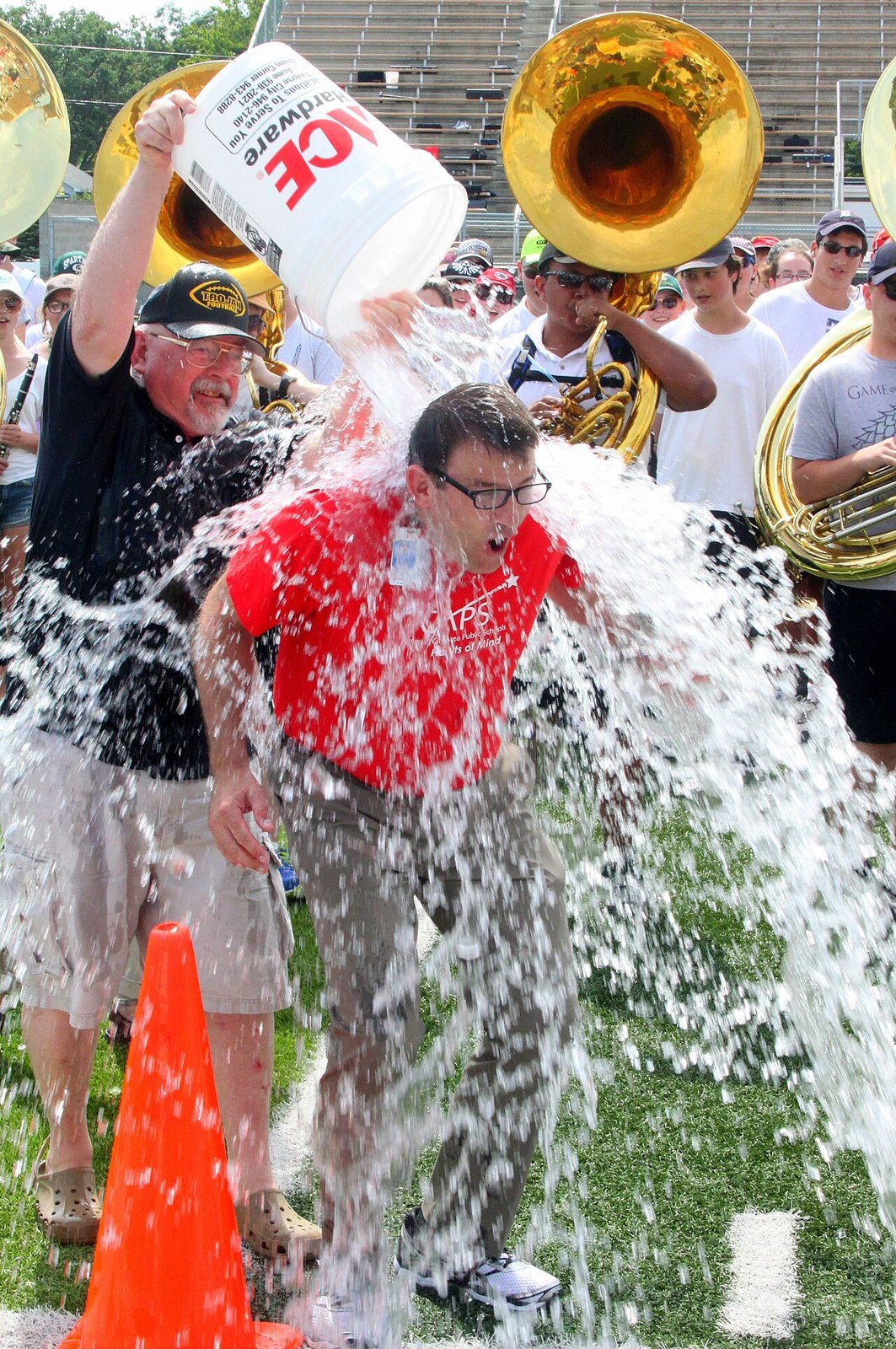 A man in a red shirt flinches as another man pours a white bucket of ice water over his head on a football field, with a marching band and tubas behind them