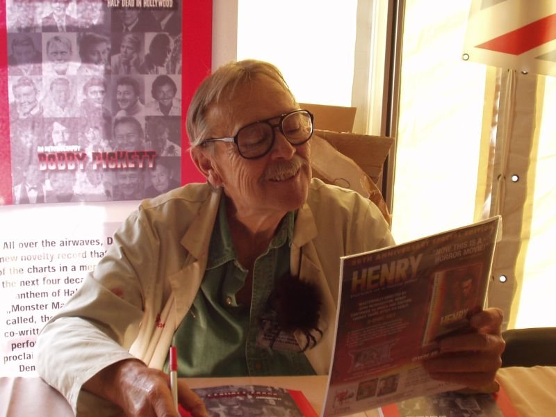 Bobby 'Boris' Pickett smiling behind a table at a fan event in 2005, holding a magazine